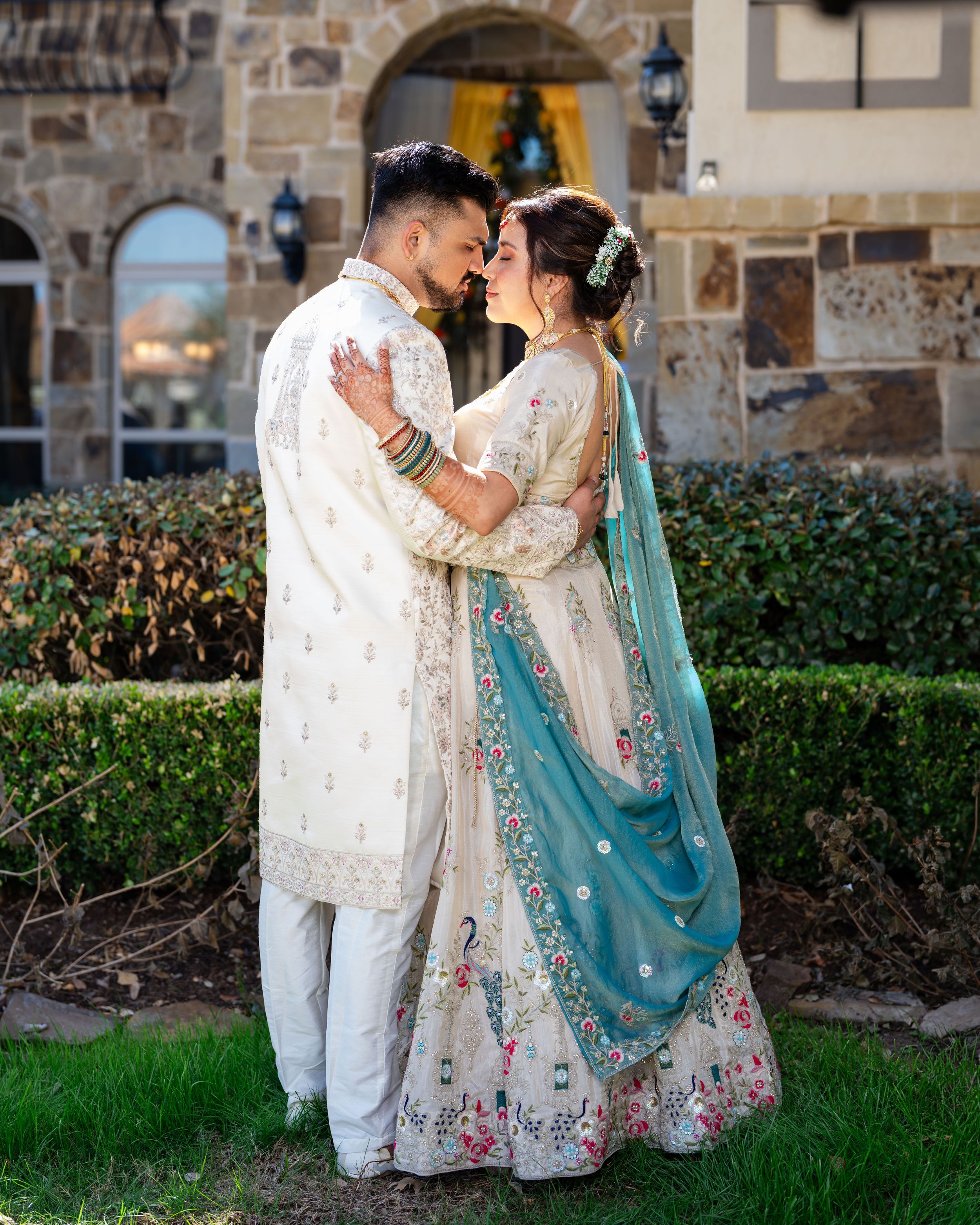 A couple in traditional South Asian wedding attire embrace outdoors against a stone wall with decorative gardens.