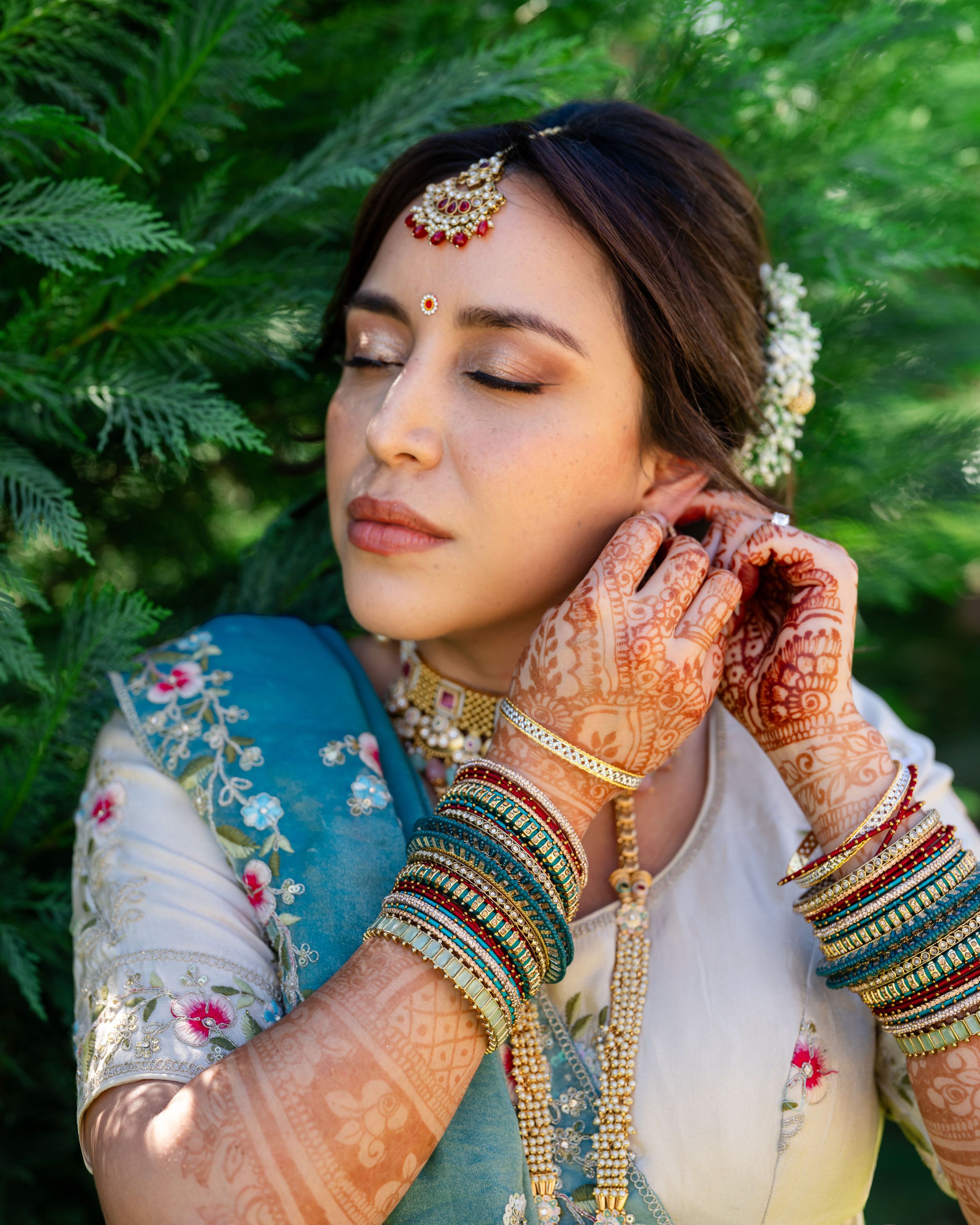 A person in traditional South Asian bridal attire with intricate henna designs and bangles poses against lush greenery.