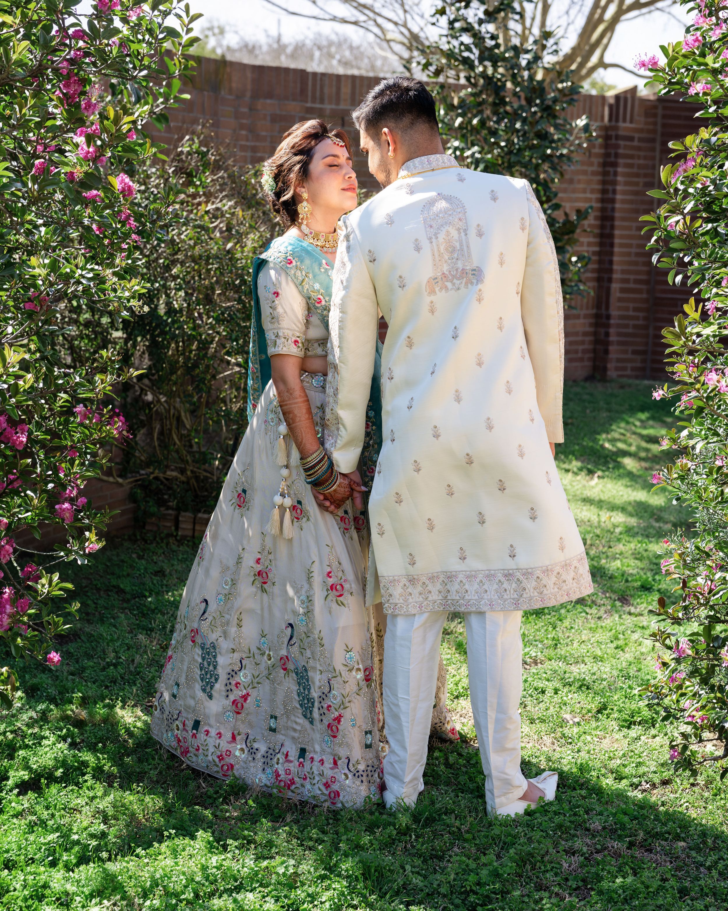 A couple in traditional South Asian attire poses together in a garden setting with pink flowers and greenery.