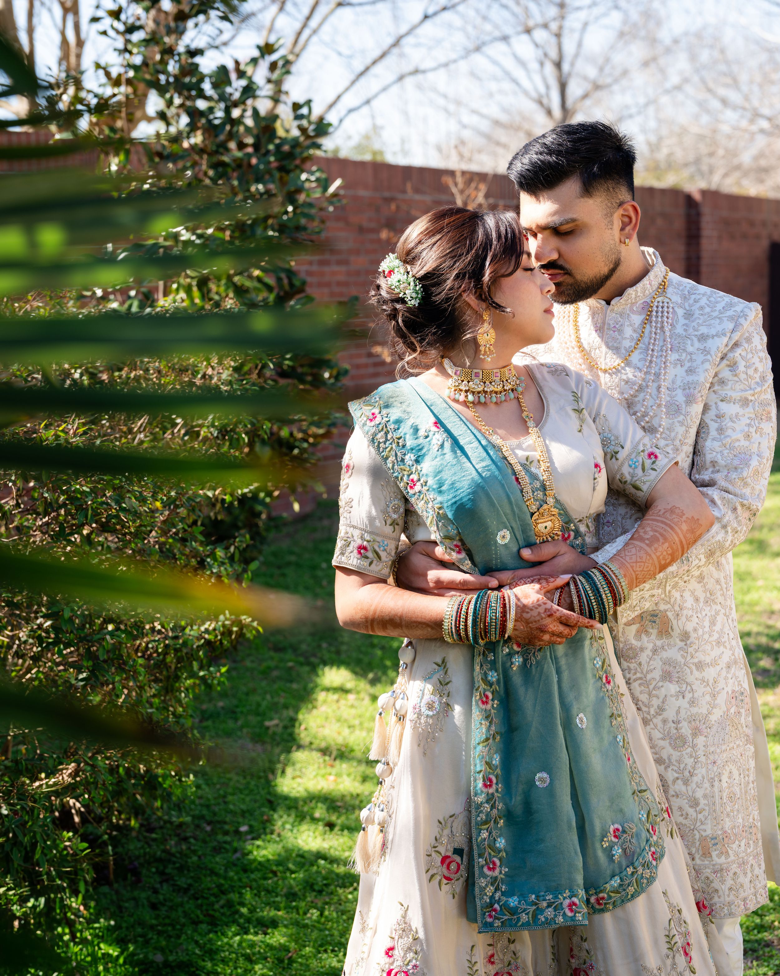 A couple in traditional South Asian wedding attire embrace in a sunlit garden setting with blooming spring trees.