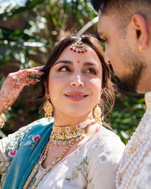A bride wearing traditional Indian jewelry and henna adorned hands smiles during a wedding ceremony outdoors.