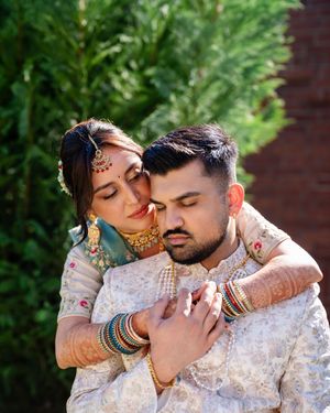 A romantic couple in traditional Indian wedding attire embraces against a green garden backdrop.