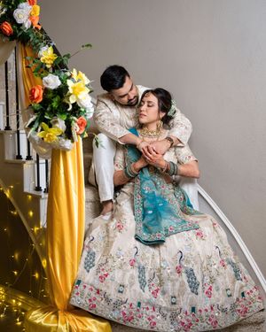 A couple in ornate Indian wedding outfits poses on stairs decorated with flowers and yellow fabric.