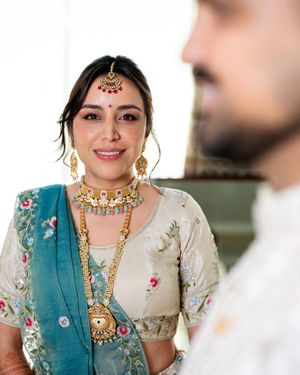 A person in traditional Indian jewelry and turquoise dupatta smiles while wearing elegant bridal attire.