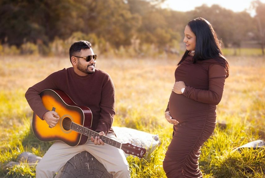 Maternity photoshoot in golden field at sunset with couple and acoustic guitar creating romantic silhouettes.