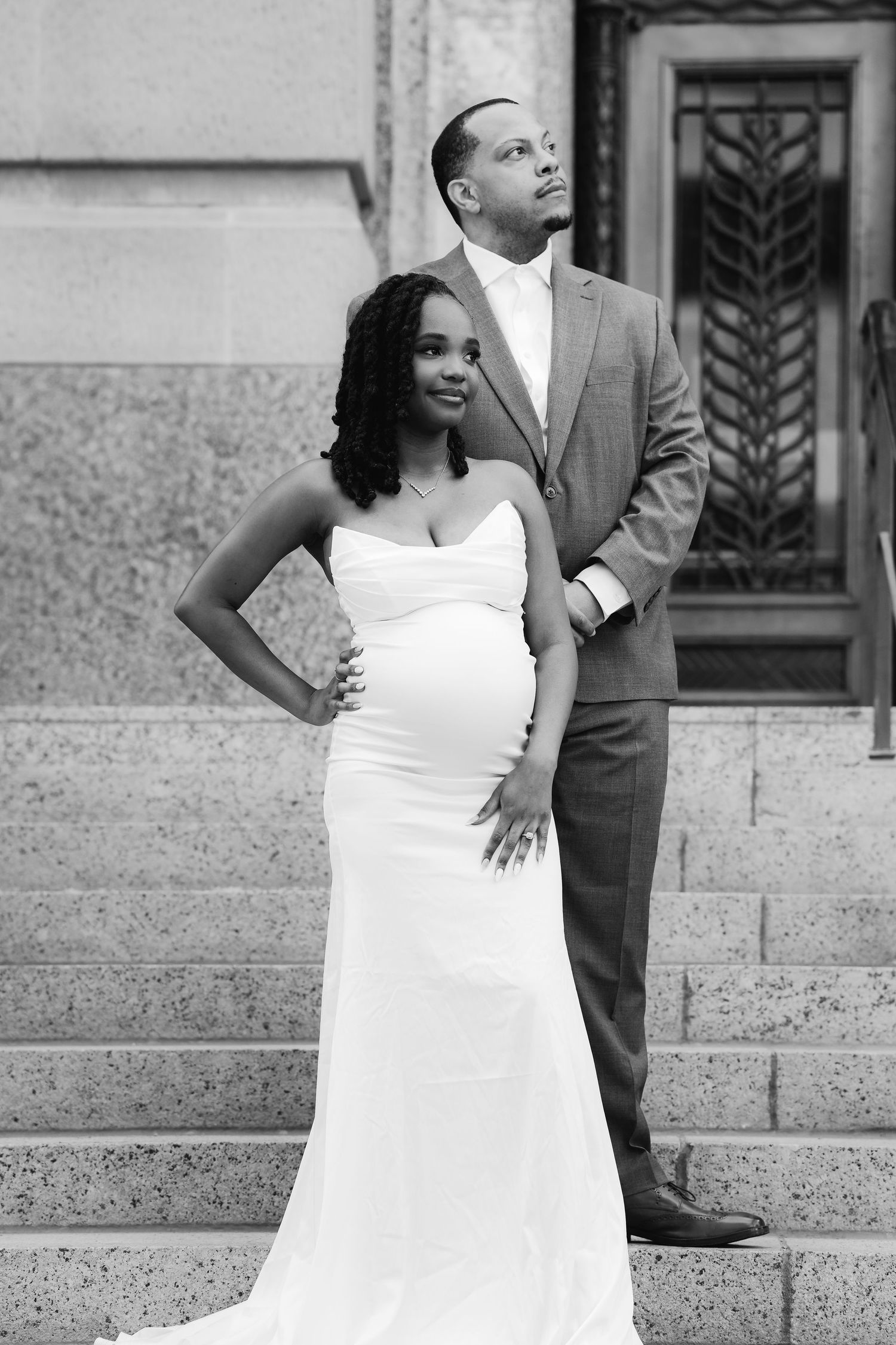 A bride in a white wedding dress poses elegantly on stone steps while her groom stands behind her.