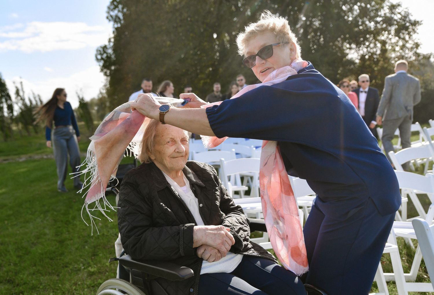 A joyful outdoor celebration with people gathered on a lawn with white folding chairs under a blue sky.
