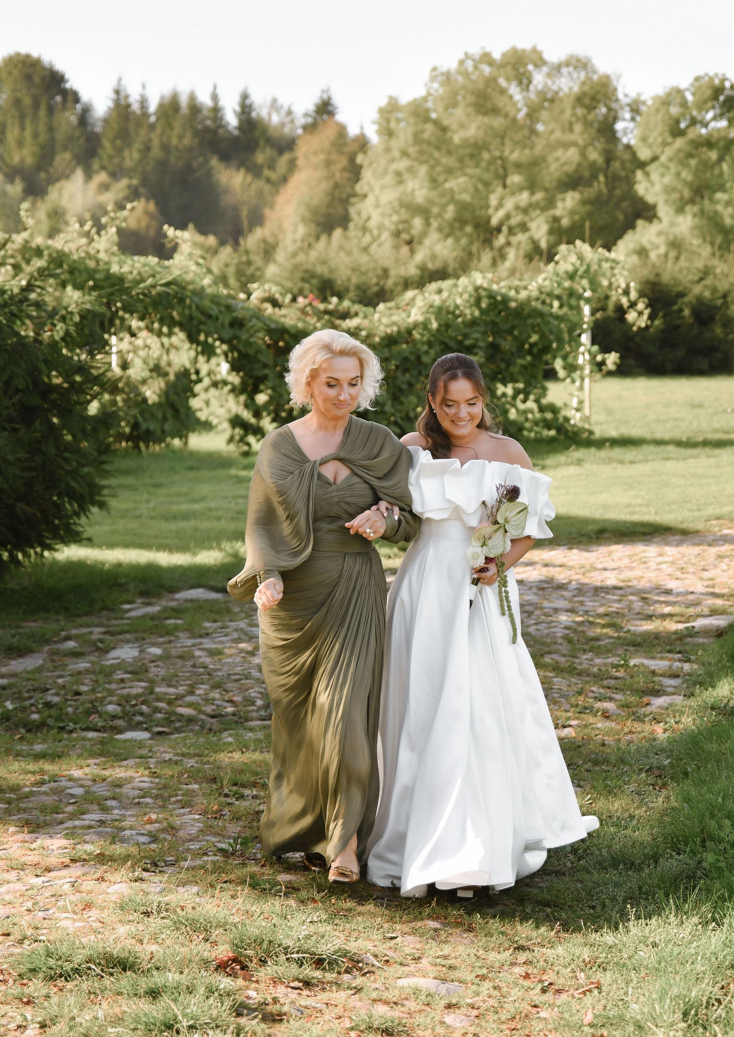 A bride in a white wedding dress walks with a companion in an olive green pleated dress through a garden setting.
