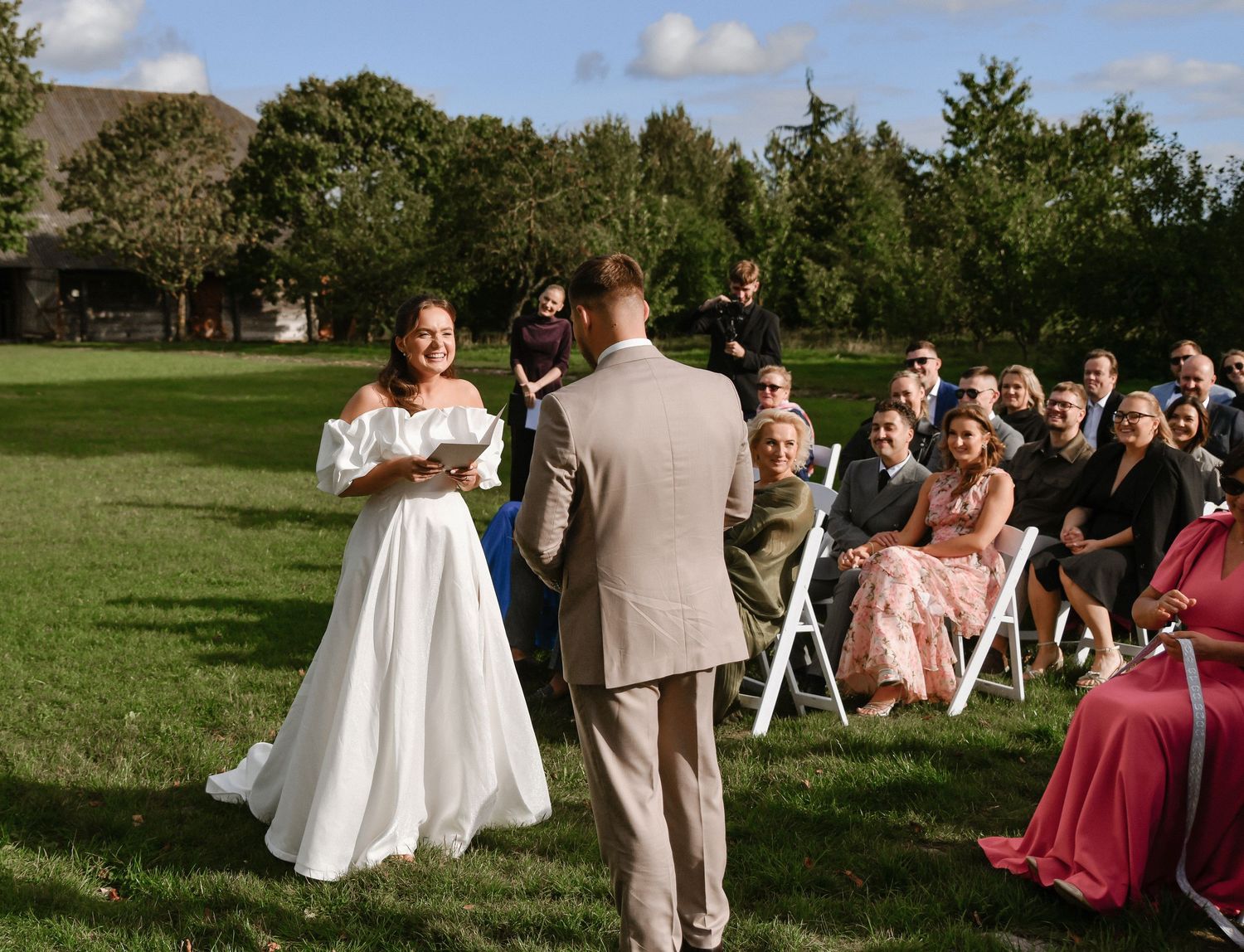 Wedding ceremony guests seated on chairs in a green outdoor garden setting during a sunny afternoon.