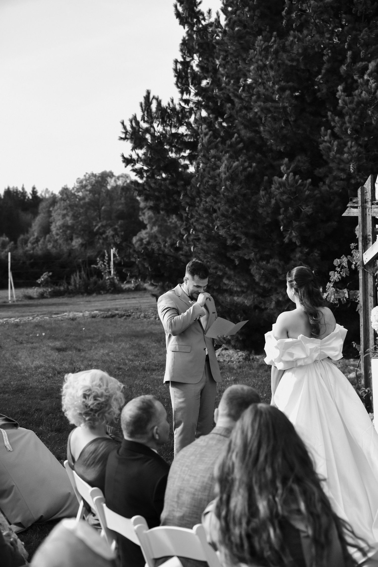 Wedding guests watch an outdoor ceremony under trees with white decor elements visible in the background.