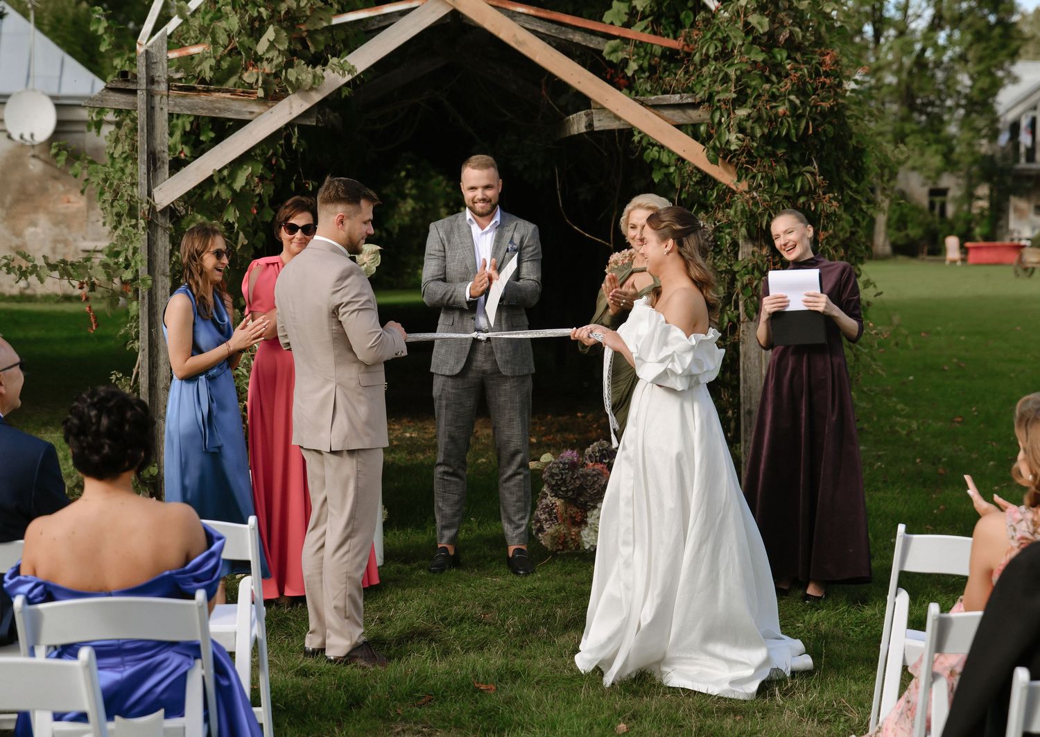 Wedding ceremony taking place under a rustic wooden arch structure in an outdoor garden setting.