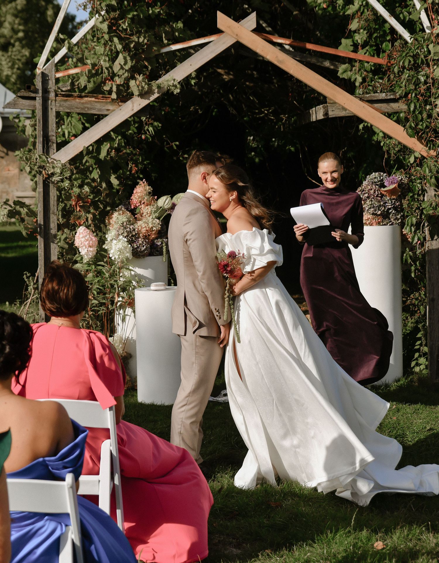 Wedding party poses for photos under wooden archway decorated with white fabric and greenery in outdoor garden setting.