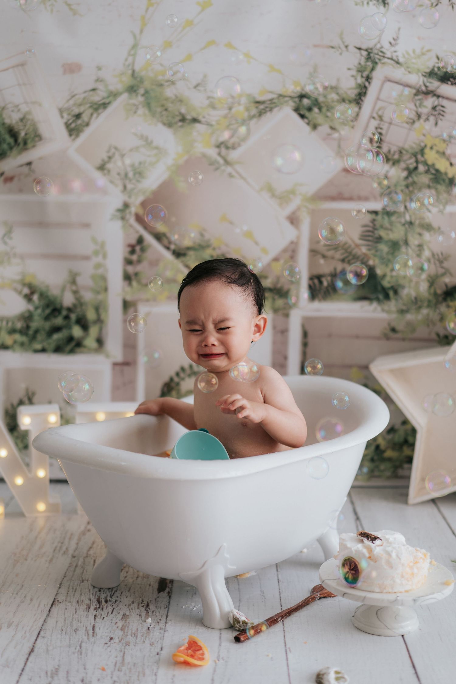 A baby enjoys a cake smash photo session in a white vintage bathtub surrounded by floral decorations.