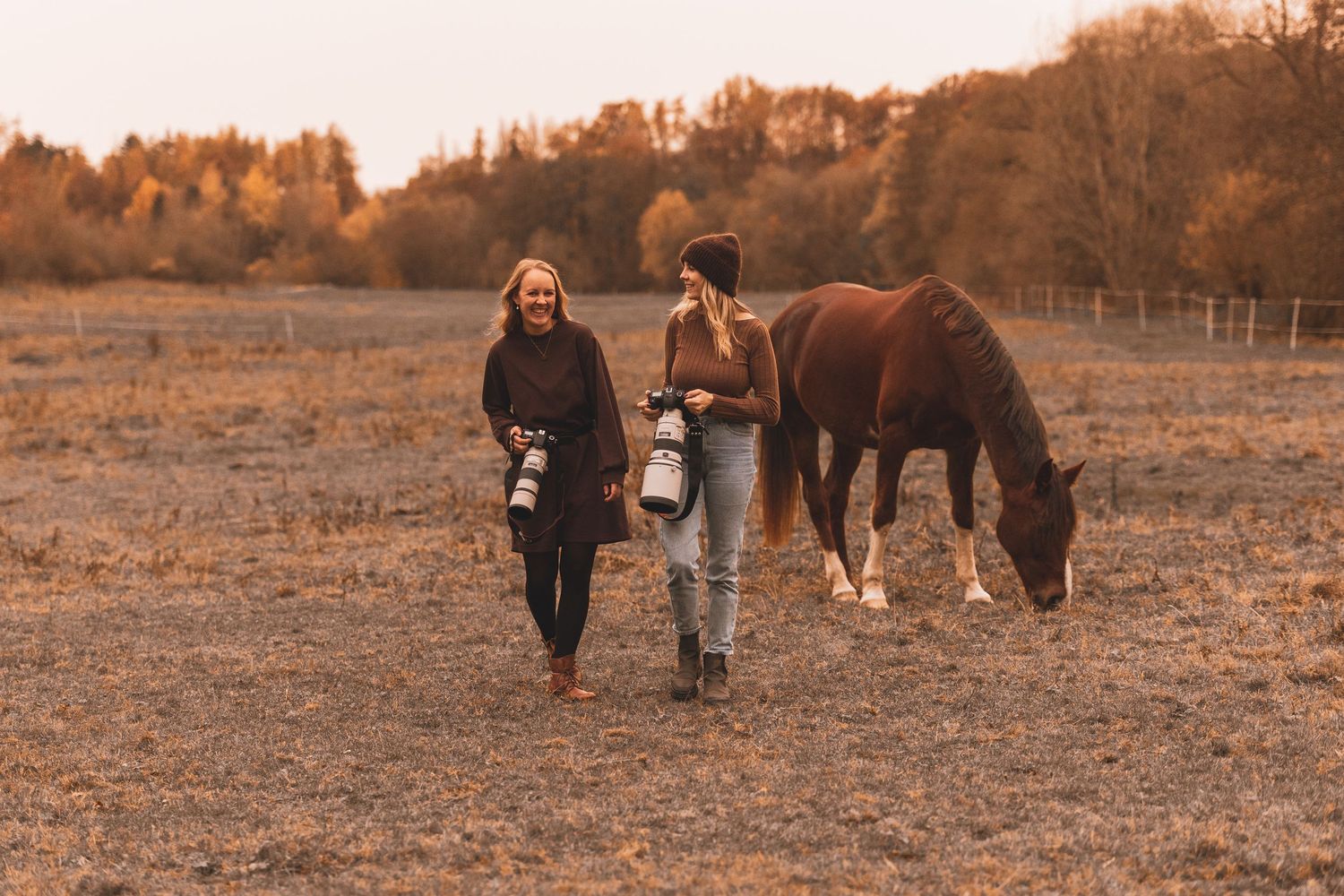 Nora Rauber Fotografie und Madlen Becker Photography gehen zusammen mit ihren Kameras in der Hand auf einer Wiese mit Pferd im Hintergrund