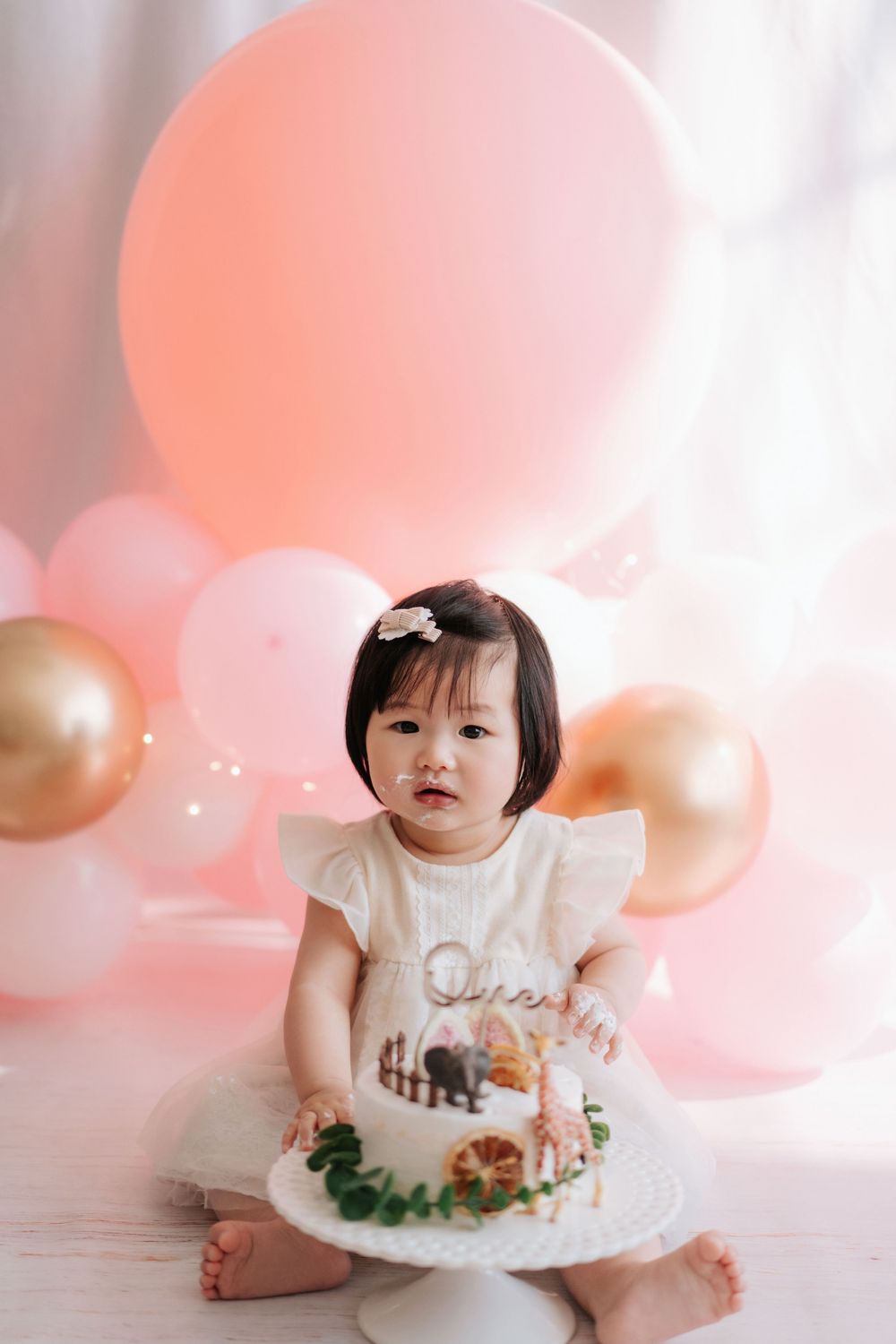 Pink and gold balloon backdrop with white cake and floral decorations for a special celebration photoshoot.