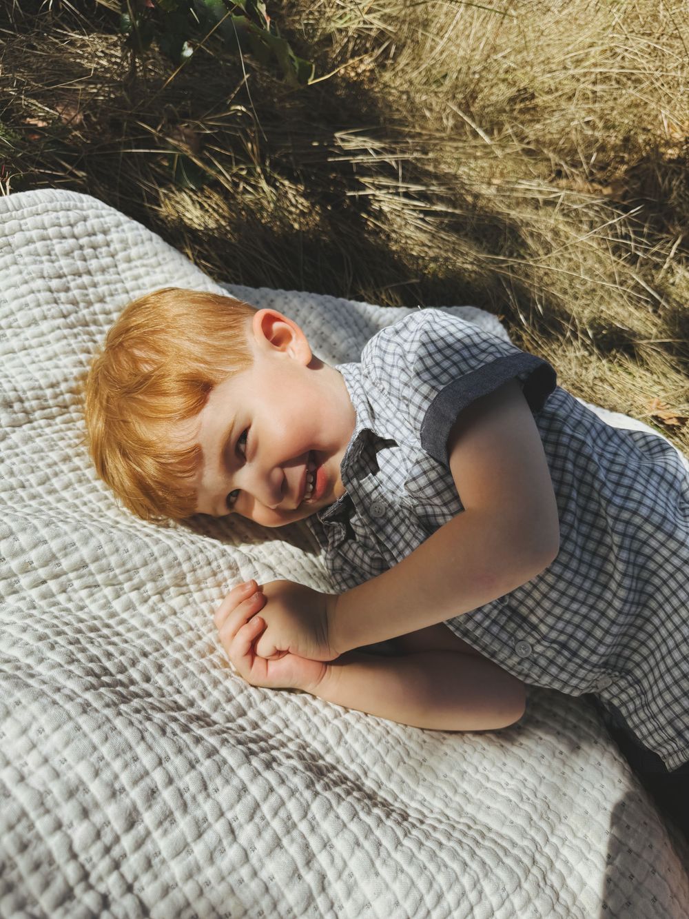 A small child in a gingham dress lies on a textured white blanket outdoors in the sunlight.