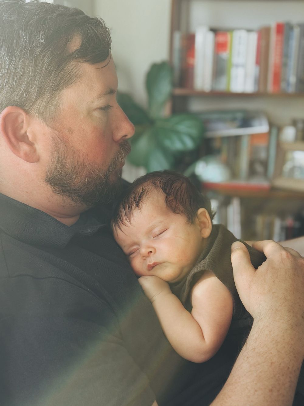 A tender moment captured as a sleeping infant rests peacefully against someone wearing black in a room with bookshelves.