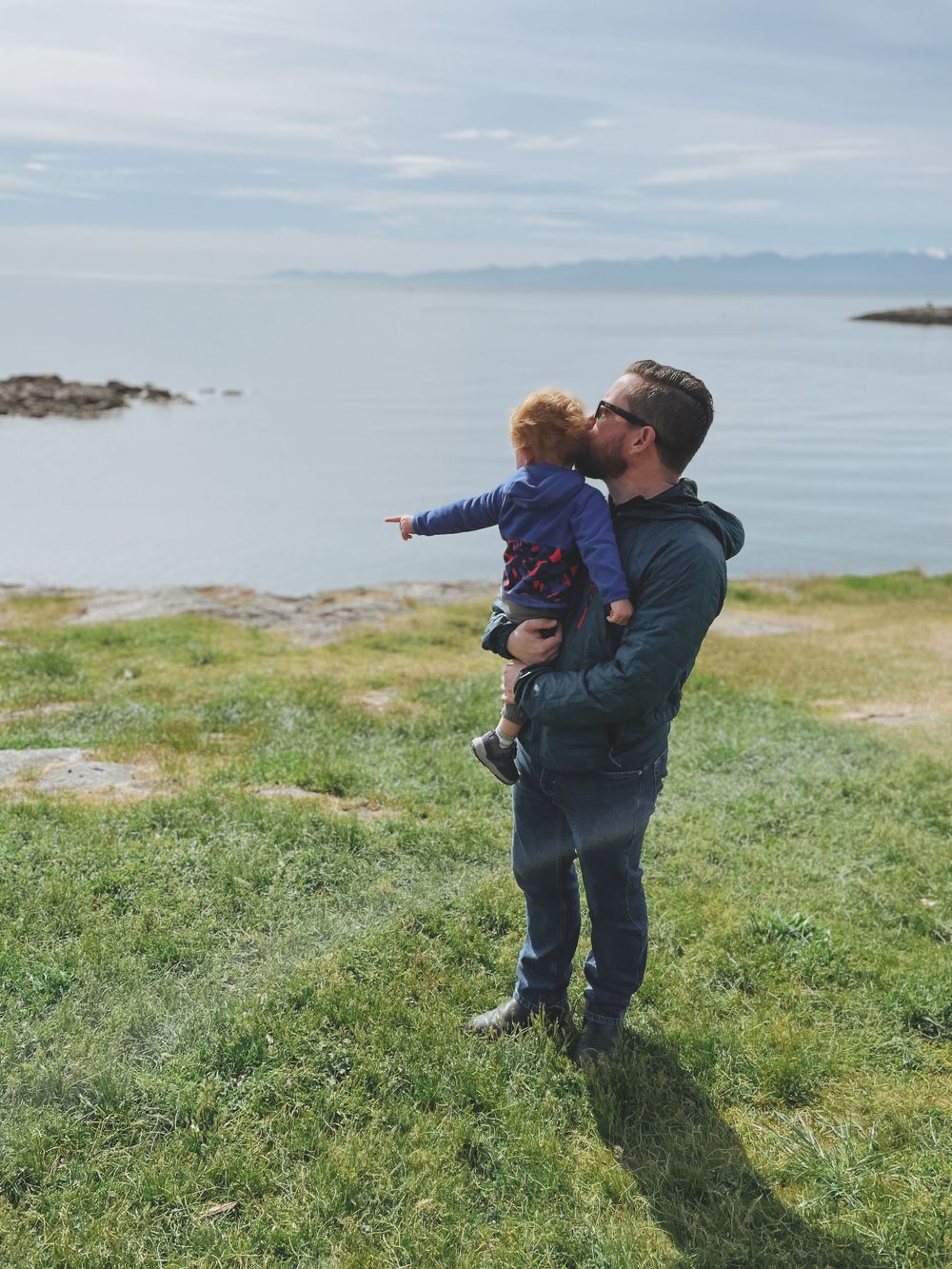 A parent and child stand together on a grassy coastal hillside overlooking a serene ocean view on a cloudy day.