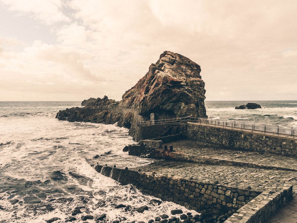 Panoramic view of a rugged coastal landscape with stone walkway and crashing waves against rocky cliffs in sepia tones.
