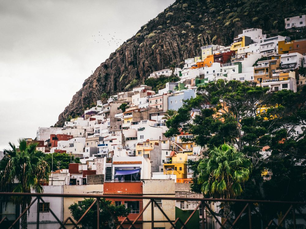 Colorful hillside houses and buildings in a coastal Mediterranean town with mountains in the background.