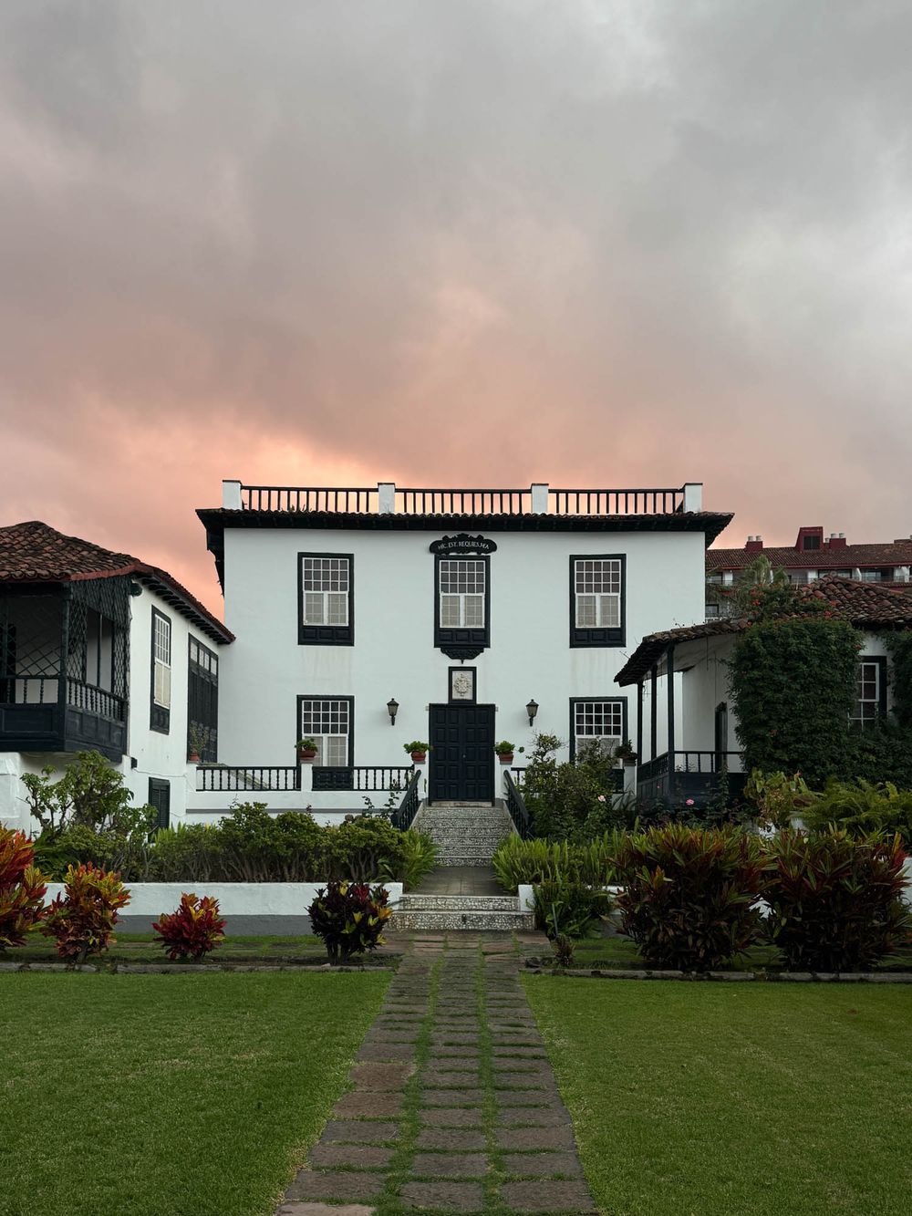 Historic white colonial mansion with black windows and symmetrical design sits on a manicured lawn under dramatic sky.