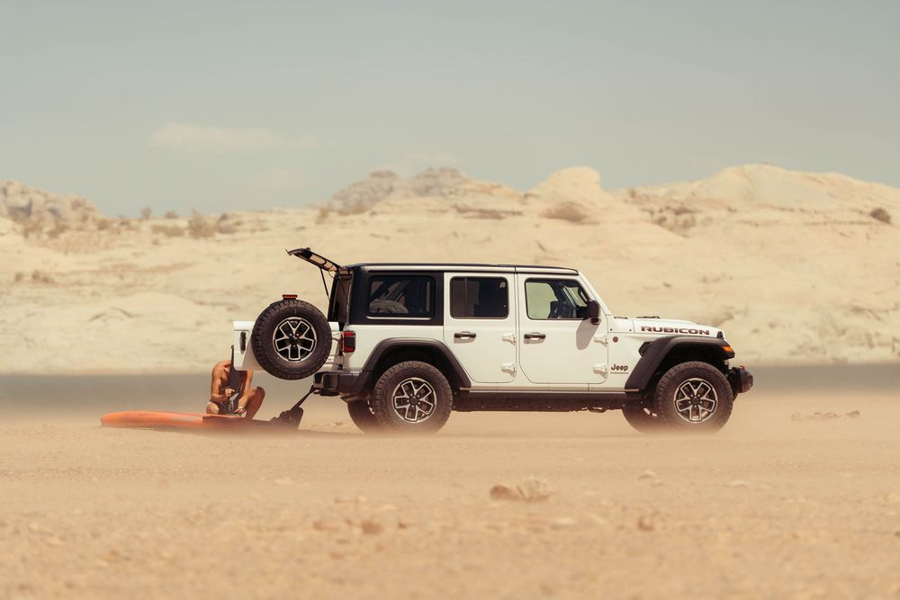 White Jeep Wrangler driving through sandy desert terrain with mountains in the background.