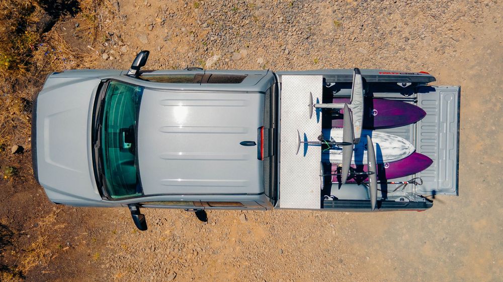 Aerial view of a silver SUV with two surfboards securely mounted on a roof rack parked on sandy terrain.