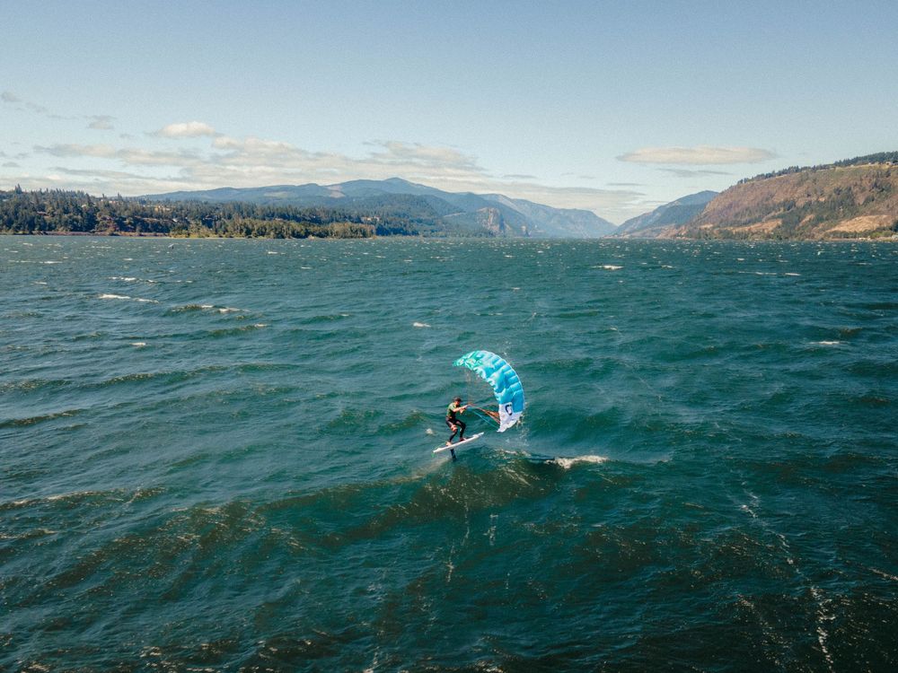 A kiteboarder rides across choppy blue-green waters with mountains visible in the distance on a sunny day.