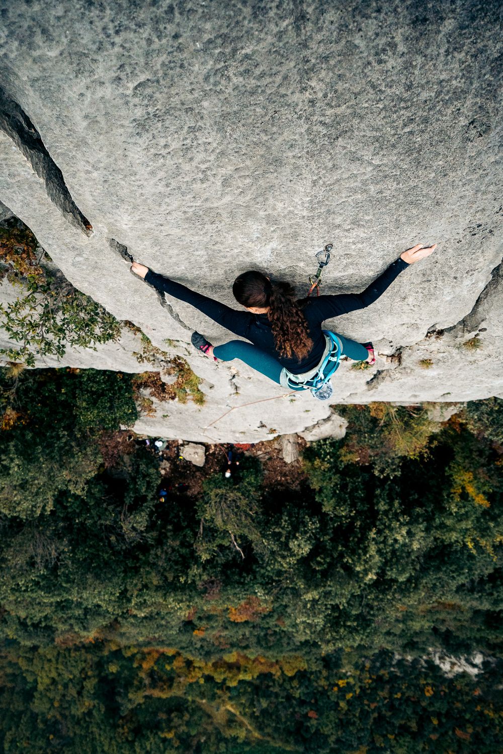 Aerial view of a rock climber ascending a steep cliff face wearing blue climbing gear against mossy rock texture.