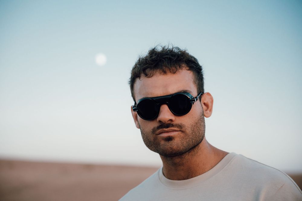A person wearing sunglasses and a black shirt poses against a bright sky in a desert landscape.