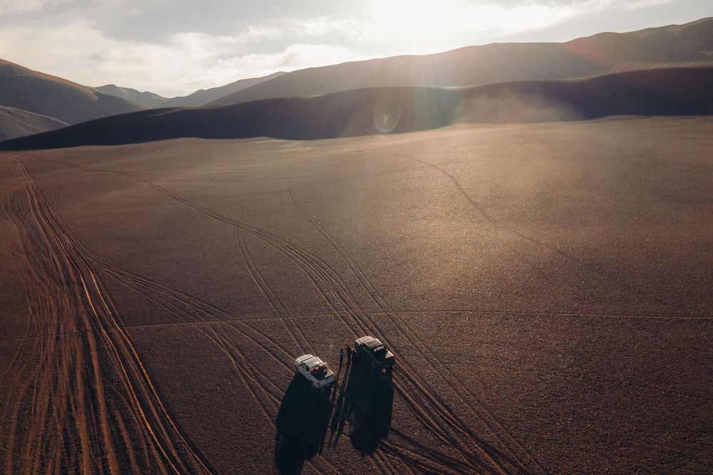 Tire tracks wind across a vast brown desert landscape at sunset with mountains silhouetted in the distance.