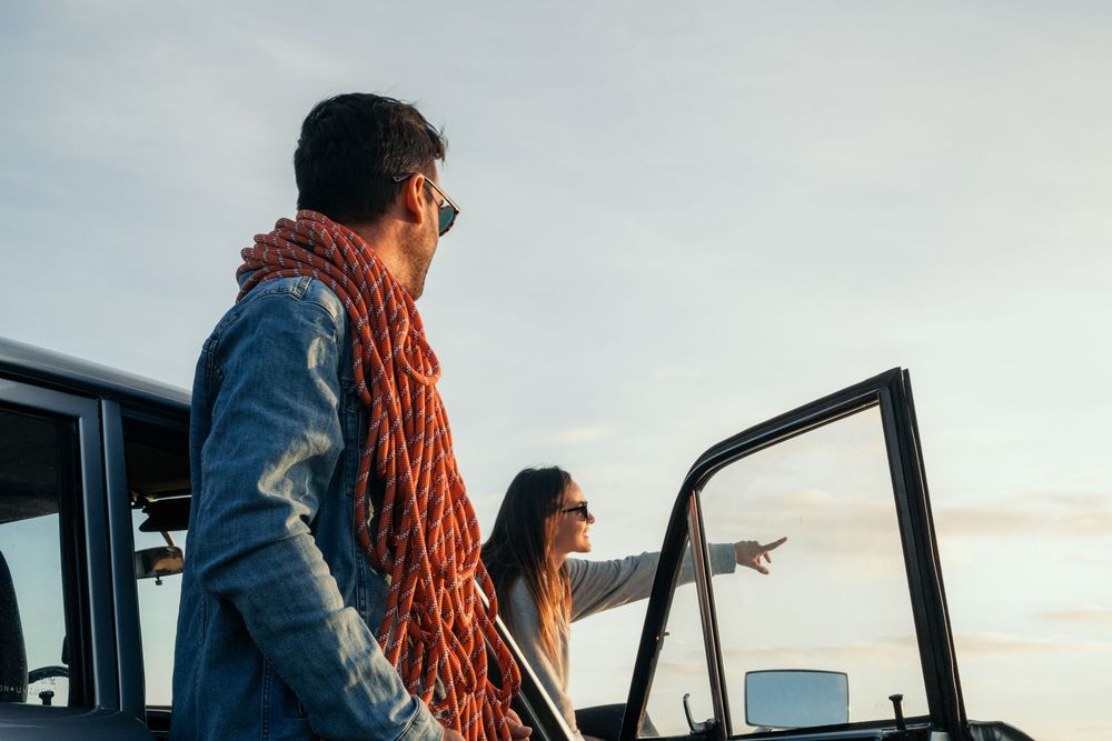 A climber wearing a blue jacket and orange rope stands at the open door of a helicopter against a cloudy sky.