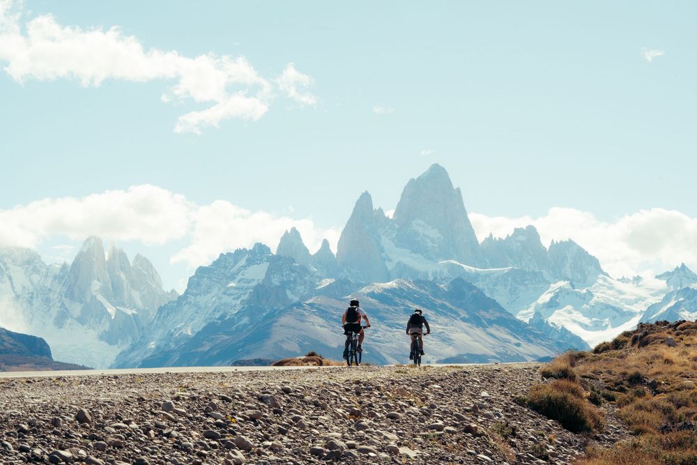 Two hikers trek along a mountain ridge with dramatic snow-capped peaks in the background against a blue sky.