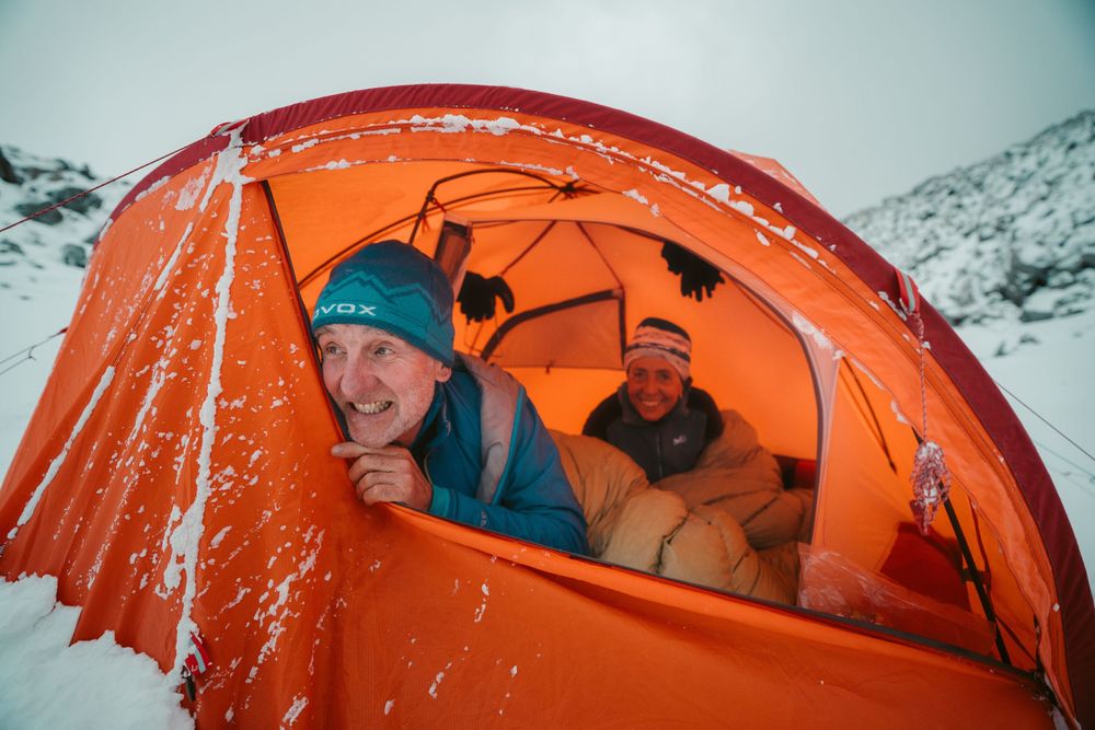 Orange mountaineering tents glow against snowy mountain landscape during winter expedition at dusk.