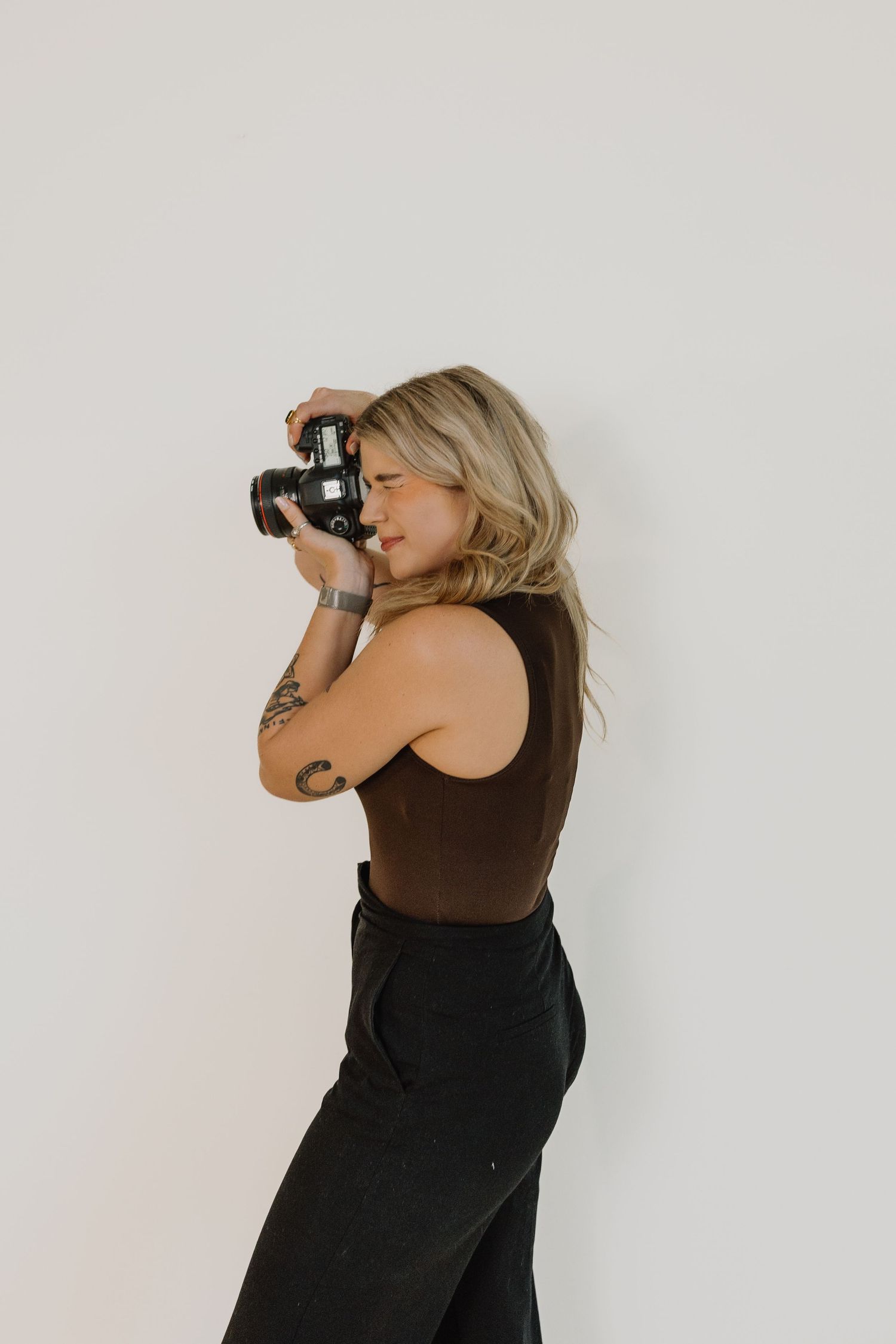Blonde woman with arm tattoo in brown sleeveless top photographs with DSLR camera against a white wall.