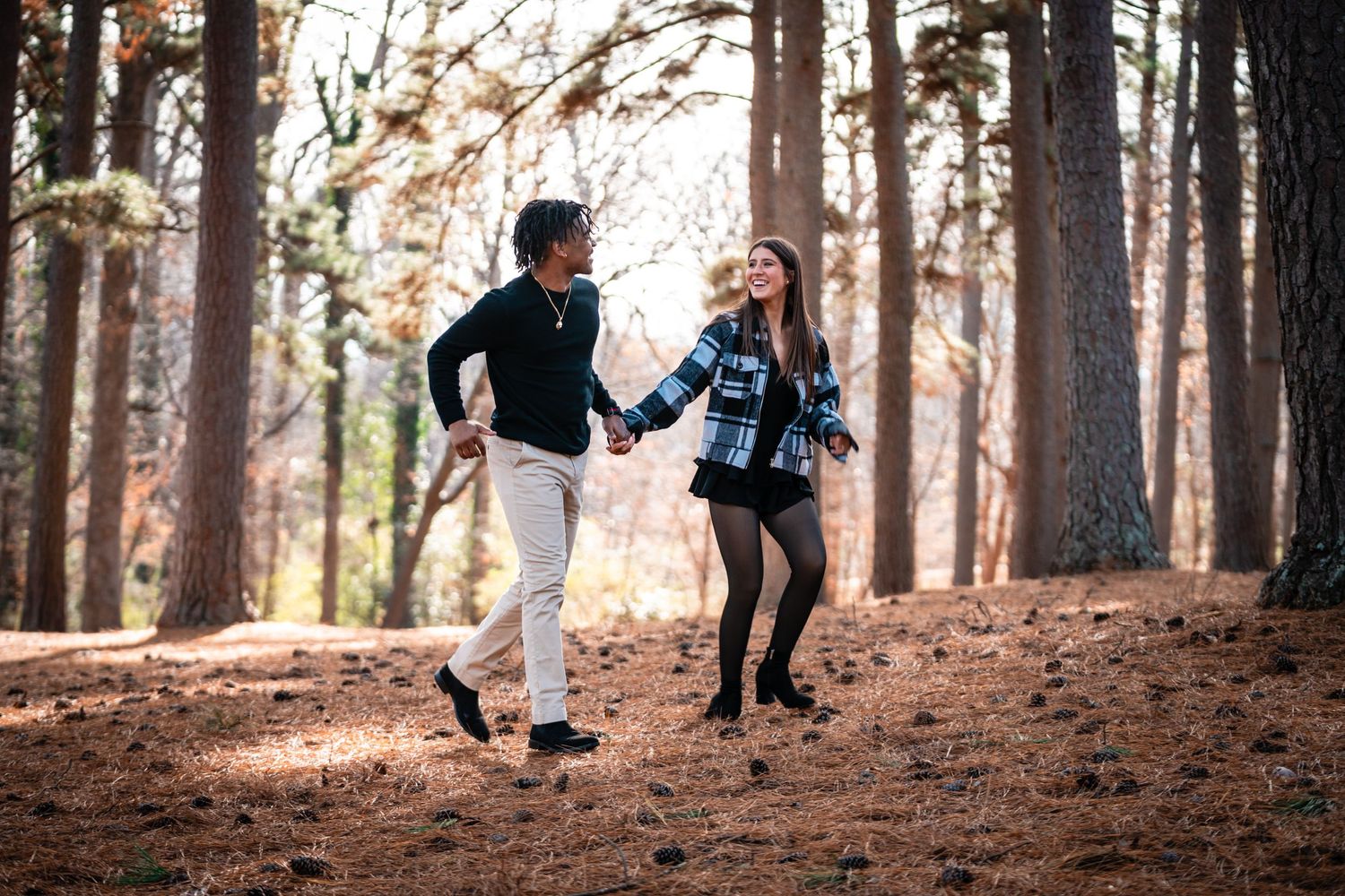 Couple holding hands while walking through a forest covered in fallen autumn leaves during golden hour.