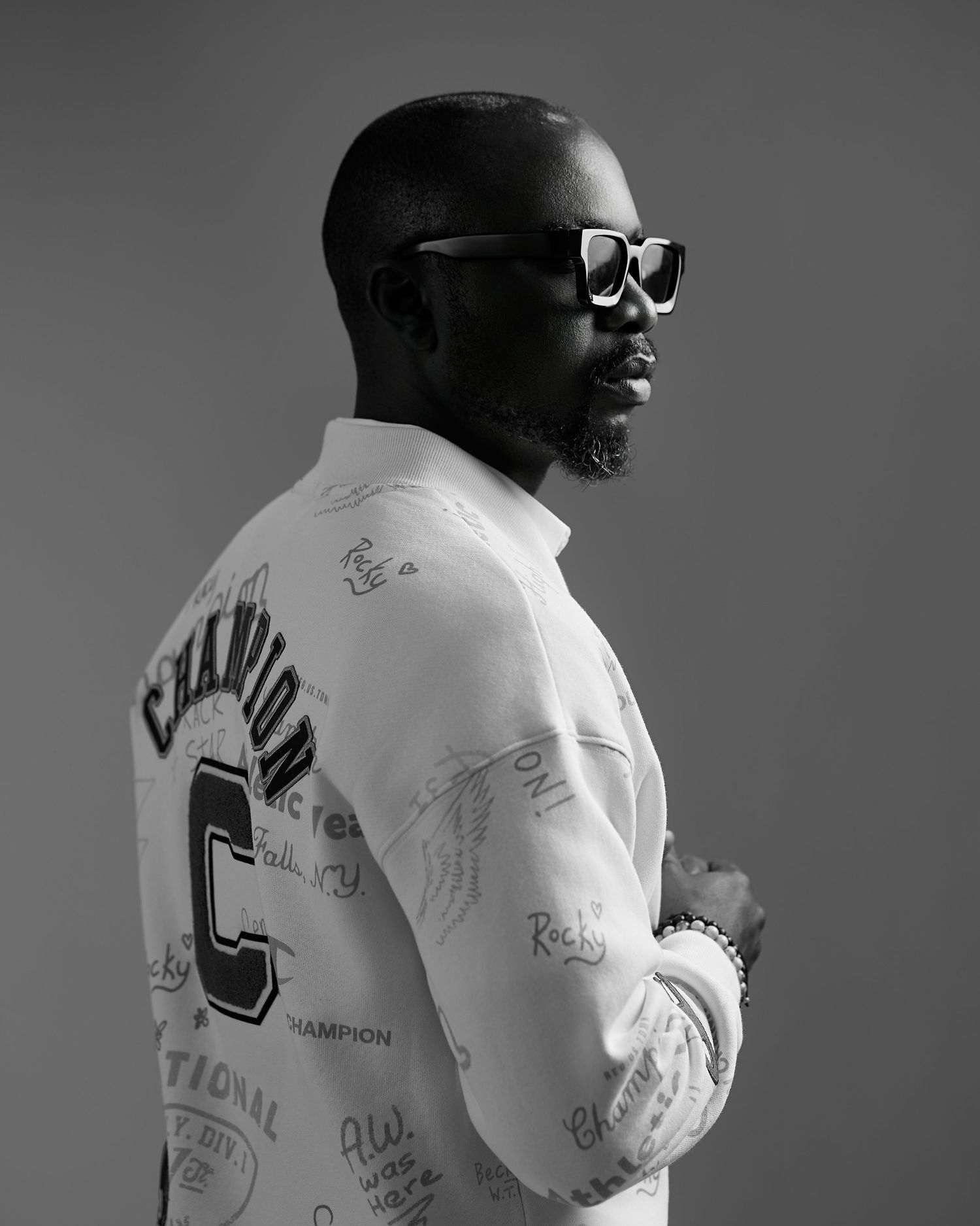 Black and white portrait of someone wearing sunglasses and a white autographed varsity-style jacket in a studio setting.