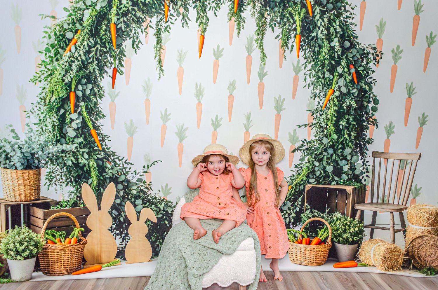 A dreamy Easter-themed photo backdrop with pastel decorations, hanging carrots, and lush greenery against a white wall.