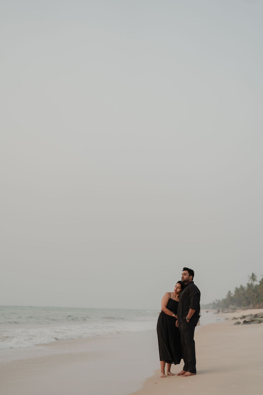 A couple walks together along a misty beach shoreline with palm trees in the distance on an overcast day.