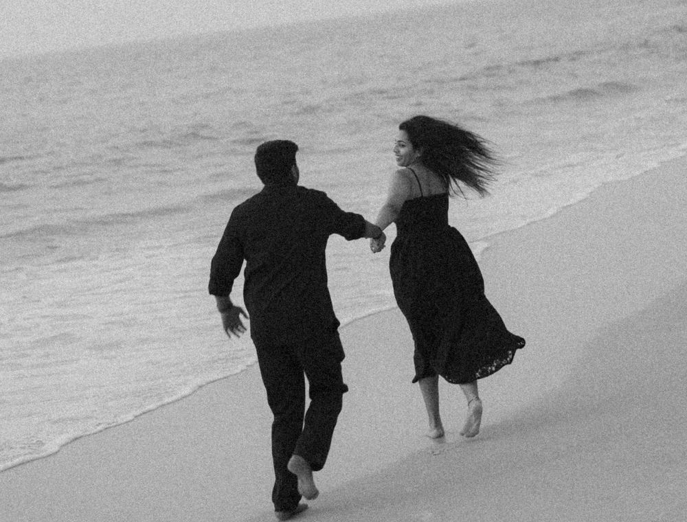 Black and white photo of a couple walking along a beach shoreline with their silhouettes reflected in the wet sand.