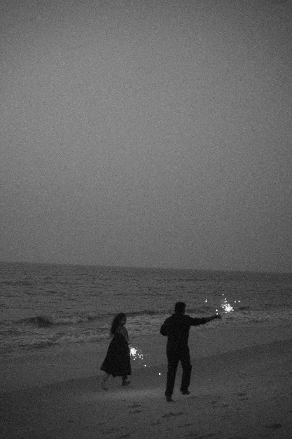 Silhouettes of people playing with sparklers on a dark beach at dusk with ocean waves in the background.