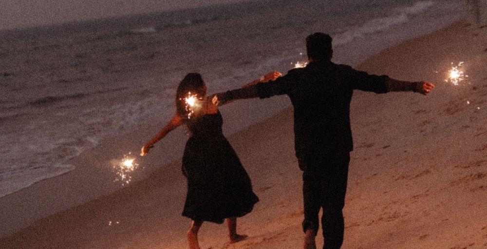 Silhouettes of people holding sparklers on a beach at dusk.