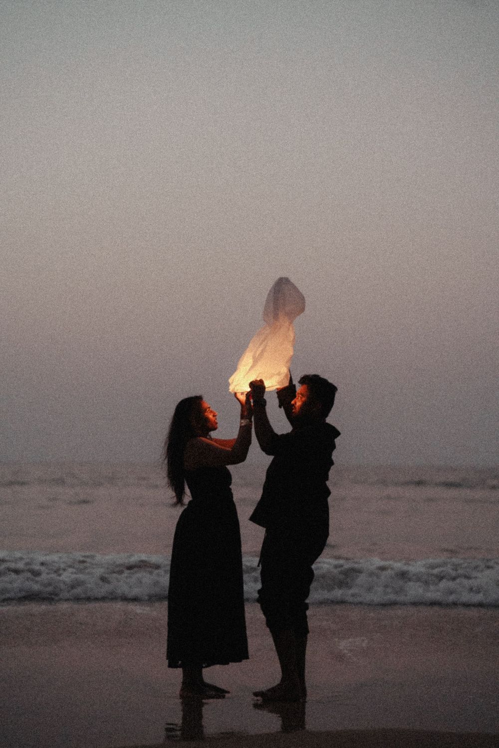 Silhouettes of two people sharing a sparkler at sunset on a beach with ocean waves in the background.