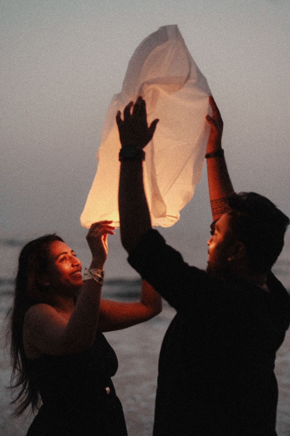 Silhouetted figures on a beach at dusk release glowing paper lanterns into the darkening sky.