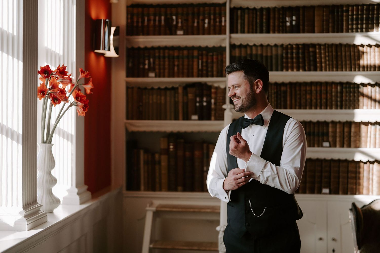 groom looking out window smiling while adjusting his cufflink in the library at Iscoyd park