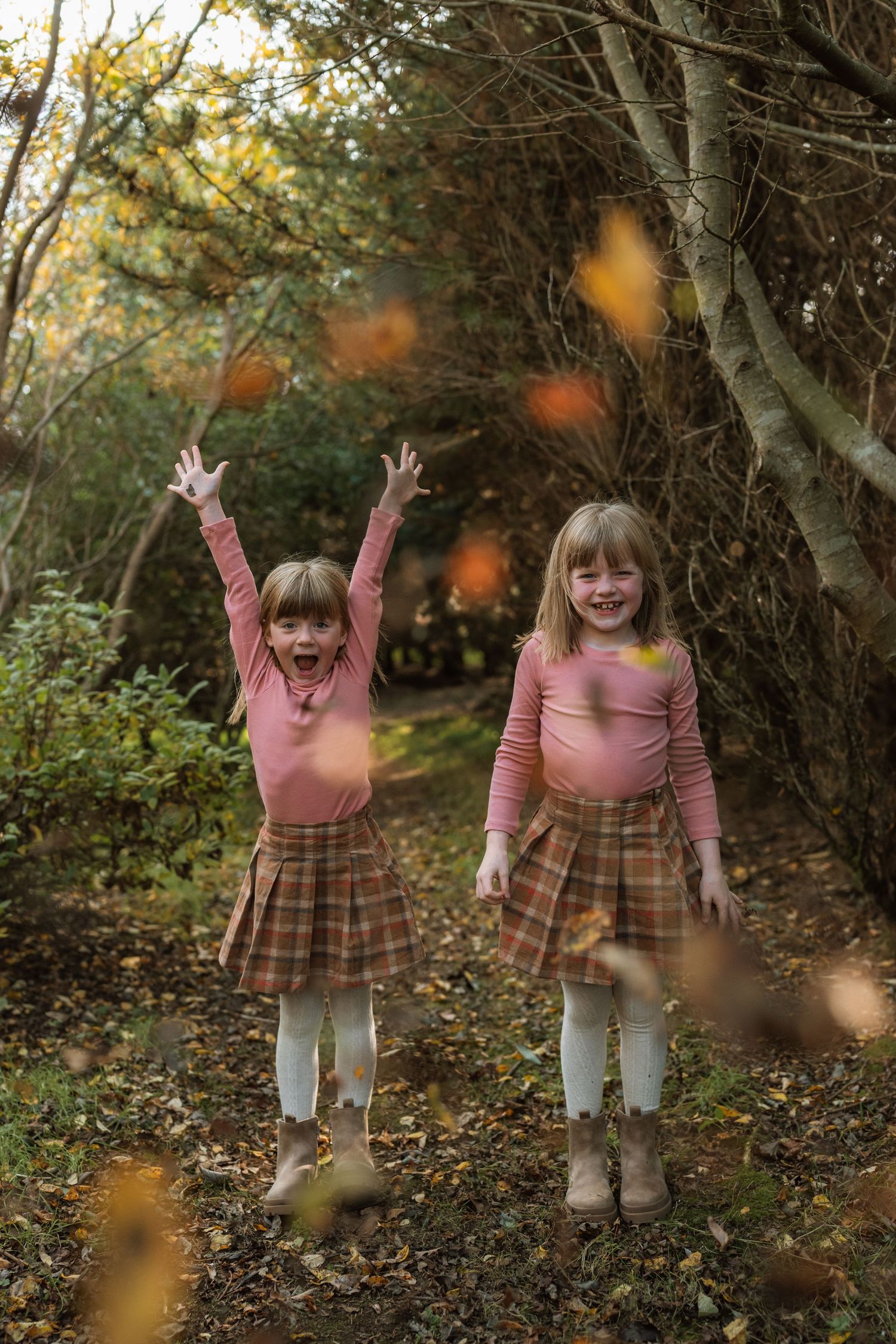 Two children in matching pink tops and plaid skirts stand together in a wooded area during autumn.