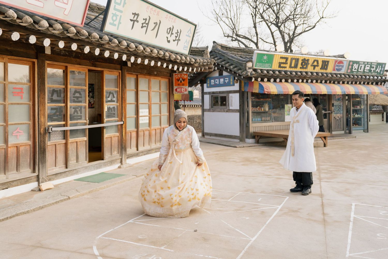 A traditional Korean hanok village street scene with people in traditional and modern clothing walking past storefronts.