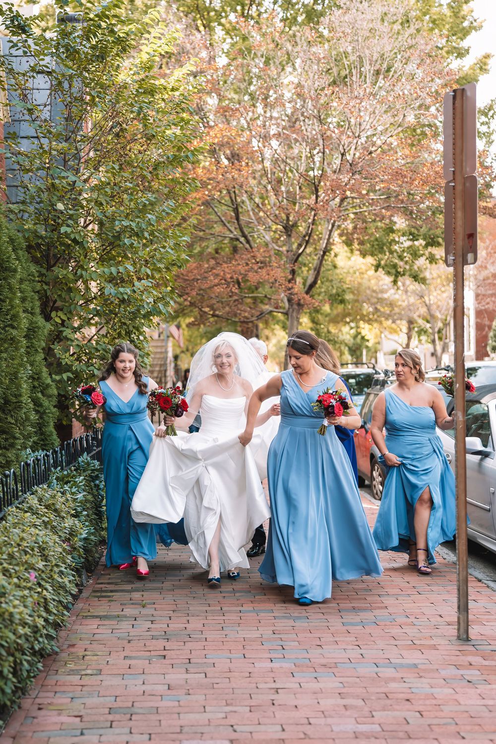 Wedding party in blue bridesmaid dresses walking along brick pathway surrounded by autumn foliage.