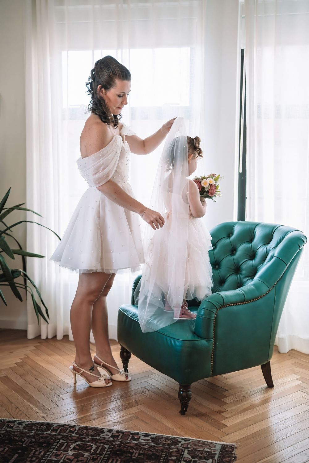 Wedding preparation scene with white dresses and a teal velvet chair in a bright room.