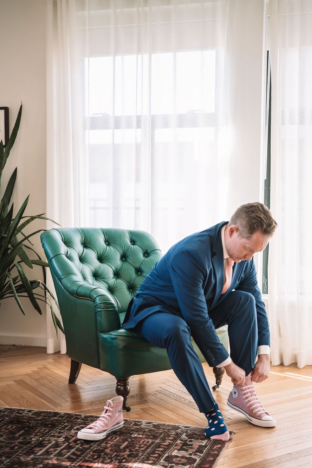 Person in navy suit putting on casual pink sneakers next to teal armchair.